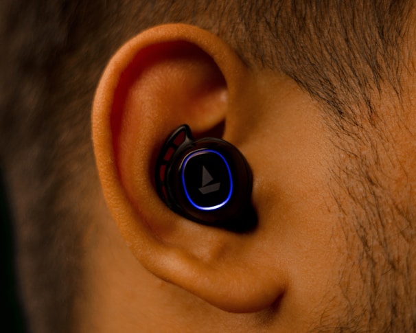 A close-up view of an ear with a black wireless earbud inserted. The earbud has a glowing blue ring around its surface, contrasting against the ear's natural skin tone.