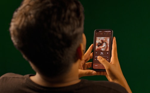 A person is holding a smartphone, looking at the music player interface displaying an album cover and song details. The person’s attention is focused on the screen, and the background is a solid green color.