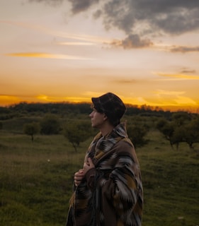 A young man wearing a hat, covered with a blanket, looking at the beautiful, orange sunset in the distance