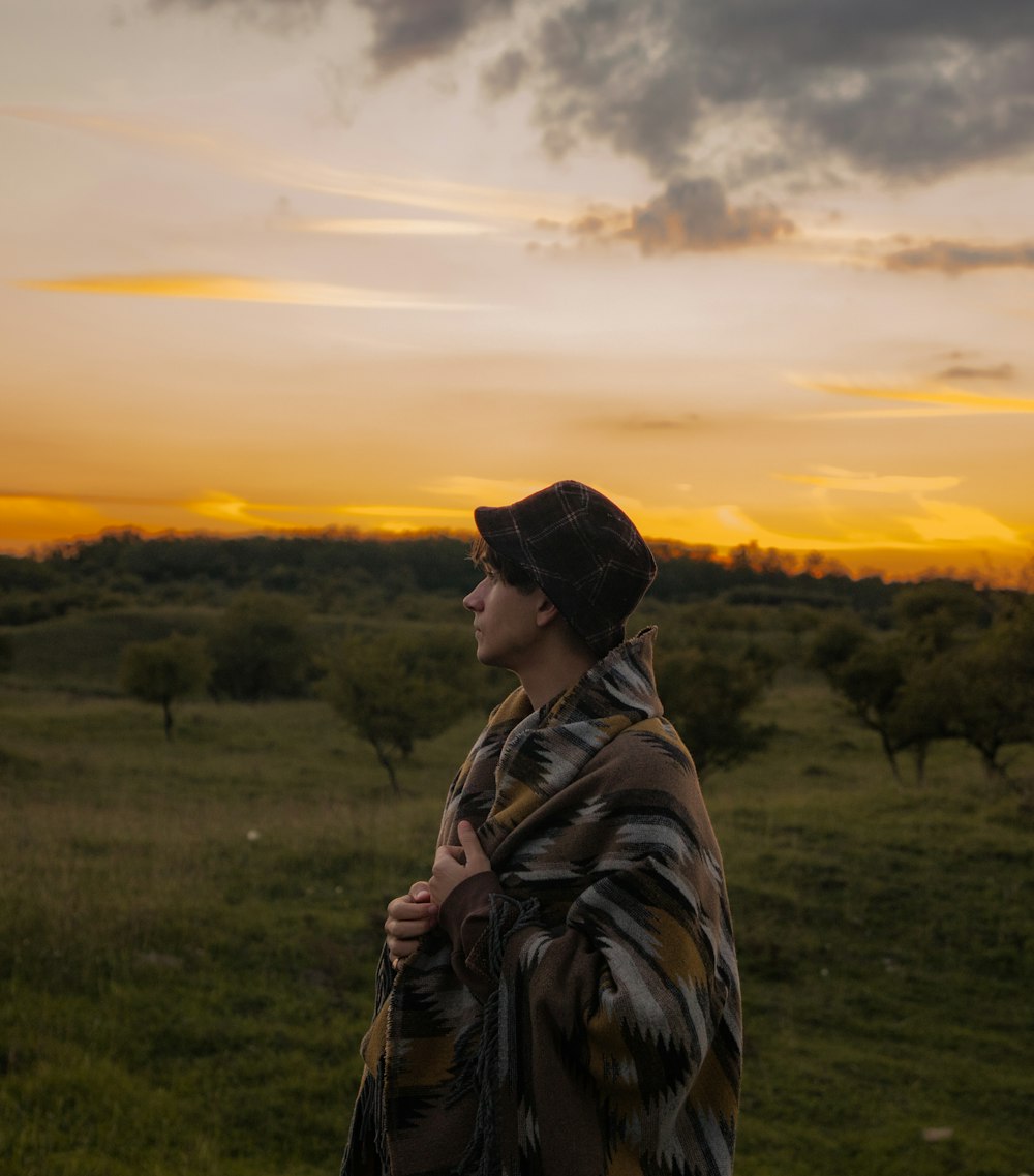 A young man wearing a hat, covered with a blanket, looking at the beautiful, orange sunset in the distance