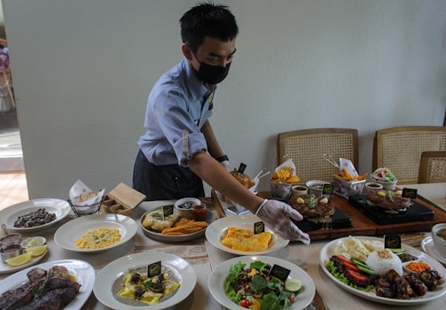 A person wearing a mask and gloves is arranging a variety of dishes on a table set for a meal. The table is filled with plates of diverse foods, including salads, meats, rice, fries, and sauces. The environment suggests a dining area with several wooden chairs.