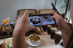 A person is taking a photo of a meal spread on a table using a smartphone. The table is set with various dishes, including a plate of food with rice, a bowl of soup, and multiple drinks with ice and lemon slices. In the background, there's a dessert and a dish with sliced mango. Another person is holding a second phone nearby.