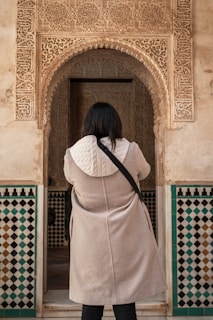A person wearing a light-colored coat with a white quilted hood stands facing an ornately decorated archway. The archway features intricate carvings and patterns, with a dominant tan and beige color. Below the arch, there are geometric patterned tiles in shades of black, white, and teal.