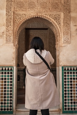 A person wearing a light-colored coat with a white quilted hood stands facing an ornately decorated archway. The archway features intricate carvings and patterns, with a dominant tan and beige color. Below the arch, there are geometric patterned tiles in shades of black, white, and teal.