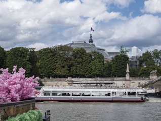 A sightseeing boat travels along a river, bordered by lush green trees and vibrant pink flowers. In the background, a prominent architectural structure with a dome is visible, along with sculptures and a waving flag on top. The sky is partly cloudy, adding a serene atmosphere to the scene.