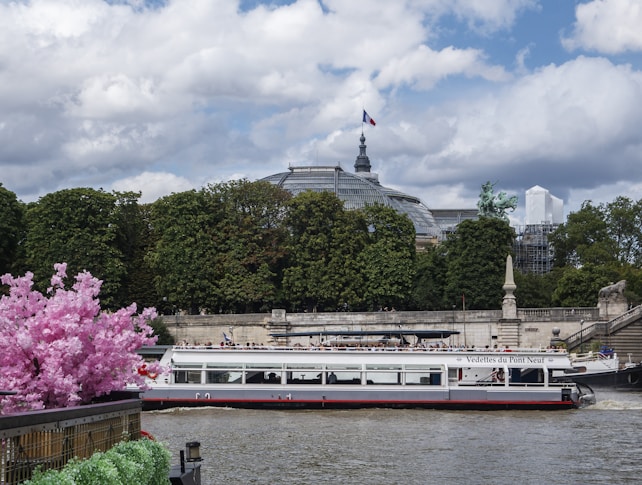 A sightseeing boat travels along a river, bordered by lush green trees and vibrant pink flowers. In the background, a prominent architectural structure with a dome is visible, along with sculptures and a waving flag on top. The sky is partly cloudy, adding a serene atmosphere to the scene.