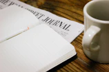 An open notebook with blank pages lying on a wooden desk beside a cup of coffee.