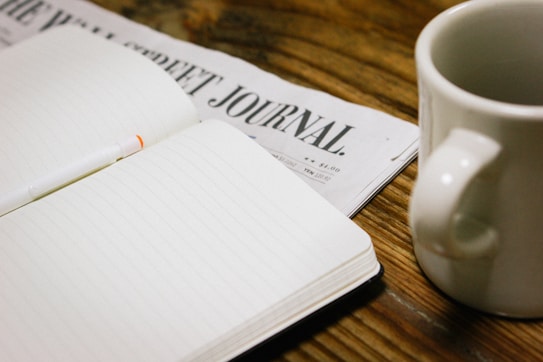 An open blank notebook with lined pages is placed on a wooden table, alongside a newspaper titled 'The Wall Street Journal' and a white coffee mug.