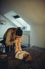 A woman leans over a young child who is lying on a bed, showing affection and playfulness. The room is softly lit, with a skylight in the background providing natural light. The setting appears warm and cozy with subdued colors.