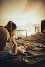 A woman is playfully interacting with a young child laying on a bed in a cozy, softly lit room. The child is smiling and dressed in a light-colored outfit, while the woman leans over, holding the child's legs gently. In the background, there is a crib, a lamp casting a warm glow, and some objects on a bedside table.
