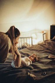 A woman is playfully interacting with a young child laying on a bed in a cozy, softly lit room. The child is smiling and dressed in a light-colored outfit, while the woman leans over, holding the child's legs gently. In the background, there is a crib, a lamp casting a warm glow, and some objects on a bedside table.