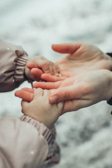 A close-up of two hands, one larger and one smaller, gently holding each other. The setting seems gentle and caring, highlighting a protective or nurturing relationship. The background is blurred, focusing attention on the hands.