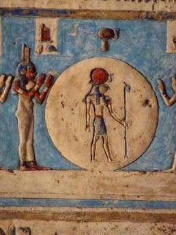 An ancient Egyptian carving depicting a deity figure holding a staff, set against a circular background. The figure is accompanied by another figure with a headpiece, both presented in traditional Egyptian attire and art style. Vibrant colors like red and blue accentuate the image, and hieroglyphic motifs decorate the surrounding space.