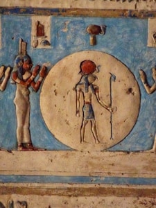 An ancient Egyptian carving depicting a deity figure holding a staff, set against a circular background. The figure is accompanied by another figure with a headpiece, both presented in traditional Egyptian attire and art style. Vibrant colors like red and blue accentuate the image, and hieroglyphic motifs decorate the surrounding space.