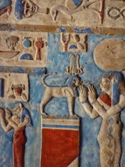 A detailed painted relief depicting figures in a classic ancient Egyptian style. Two figures are shown in profile, adorned with traditional Egyptian garments and headdresses. The scene includes a large cat-like animal perched on a rectangular platform. Hieroglyphs and additional symbols fill the background, intricately carved and painted with vibrant colors.