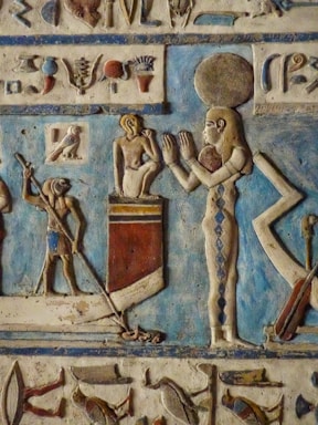 A detailed ancient Egyptian relief depicting multiple figures, including a standing figure with an ornate headdress and a smaller figure on a boat. The background is predominantly blue with hieroglyphs and symbols above the figures. The art is carved with intricate details and vibrant colors.