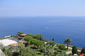 A lush green garden with various trees and shrubs overlooks a vast, calm blue ocean. The garden features palm trees and a structure with a pergola, surrounded by dense greenery. Sun lounges are visible to the left side of the garden, and a small white boat can be seen floating on the ocean in the distance.