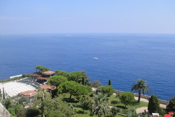A lush green garden with various trees and shrubs overlooks a vast, calm blue ocean. The garden features palm trees and a structure with a pergola, surrounded by dense greenery. Sun lounges are visible to the left side of the garden, and a small white boat can be seen floating on the ocean in the distance.
