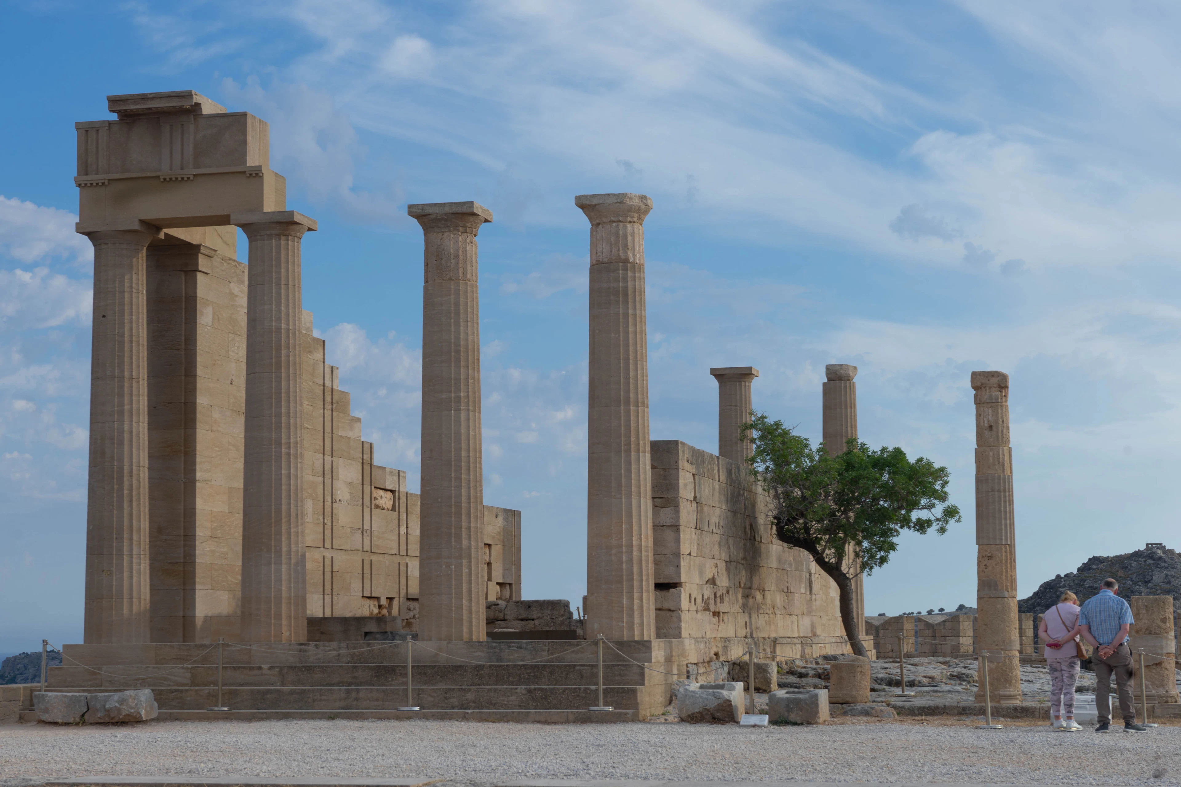 The standing Doric columns of the ancient temple complex on the Acropolis of Lindos, Rhodes, with visitors and a lone tree beside the weathered stonework under a bright sky.