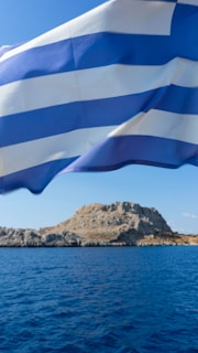 A flag with alternating blue and white horizontal stripes is prominently displayed in the foreground, billowing gently in the wind. In the background, a rugged, rocky island is surrounded by calm, deep blue sea under a bright, clear sky.