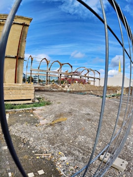 A construction site with a partially built, rusted steel structure in the background. The ground is covered with gravel, dirt, and construction debris. A stack of wooden boards is located on the left. A fenced barrier is observable in the foreground, while a building with a tall, yellow spire and a weather vane is seen in the distance under a clear blue sky.