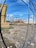 A construction site with a partially built, rusted steel structure in the background. The ground is covered with gravel, dirt, and construction debris. A stack of wooden boards is located on the left. A fenced barrier is observable in the foreground, while a building with a tall, yellow spire and a weather vane is seen in the distance under a clear blue sky.