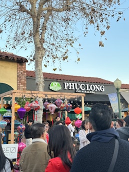 A bustling scene outside a Phuc Long Coffee & Tea shop, featuring a gathering of people engaged in conversation and observation. The storefront is decorated with colorful lanterns hanging from a wooden frame, adding to the lively atmosphere. A bare tree is visible in the background against a clear sky.