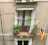 A small balcony with potted plants and a hanging colorful flag. The balcony features a wooden door with curtains. There are clotheslines with a yellow clothespin attached.
