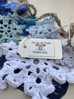 A collection of crocheted snowflakes in various shades of blue and white is spread on a surface. They are accompanied by a rustic twine and a tag labeled 'Jill-Of-All-Trades Crocheted Goods'.