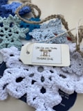 A collection of crocheted snowflakes in various shades of blue and white is spread on a surface. They are accompanied by a rustic twine and a tag labeled 'Jill-Of-All-Trades Crocheted Goods'.