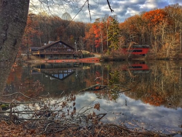 A serene lakeside scene featuring a rustic cabin on the left, surrounded by autumn-colored trees. The clear water reflects both the cabin and the vivid orange and red foliage, creating a mirrored image. A covered bridge is visible in the background, adding to the picturesque landscape. Bare branches and dried leaves can be seen in the foreground, adding to the seasonal ambiance.