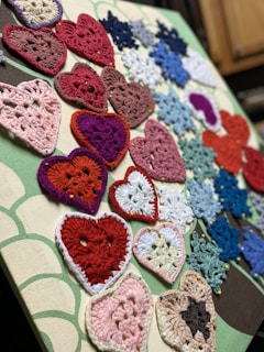 A collection of crocheted hearts and snowflakes pinned onto a fabric-covered board. The hearts are made in various colors like red, pink, and purple, while the snowflakes are mostly in shades of blue and white. They are arranged in an overlapping pattern, displaying a variety of crochet designs.