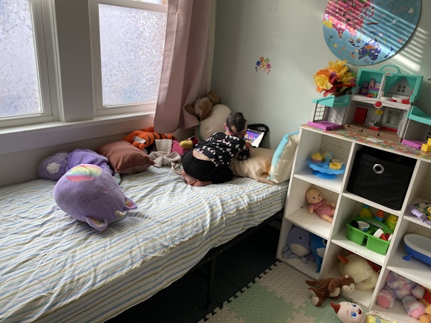 A child is positioned on a bed, focused on a tablet. The bed has a striped bedsheet with several stuffed animals, including a large purple unicorn pillow. A shelving unit is visible next to the bed, containing a toy house, a doll, and various other toys. The room is softly lit by natural light coming through the window, which is partially covered by pink curtains.