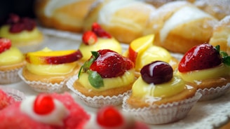 A selection of colorful pastries decorated with fresh fruits like strawberries, peaches, and cherries, topped with creamy custard in paper liners. In the background, there are bread rolls partially covered with powdered sugar.