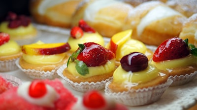 A selection of colorful pastries decorated with fresh fruits like strawberries, peaches, and cherries, topped with creamy custard in paper liners. In the background, there are bread rolls partially covered with powdered sugar.