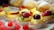 A selection of colorful pastries decorated with fresh fruits like strawberries, peaches, and cherries, topped with creamy custard in paper liners. In the background, there are bread rolls partially covered with powdered sugar.