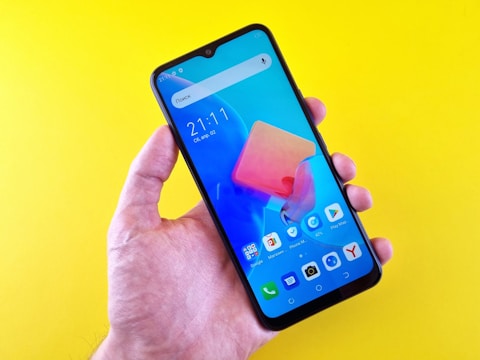 A hand holding a smartphone with a vibrant display showing various app icons, against a bright yellow background. The screen displays the time, date, and a search bar.