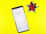 A smartphone displaying its system information screen is placed on a bright yellow surface. To the right of the smartphone, there is a patch or sticker featuring a black and white star outline with a red hand making a rock-and-roll sign in the center.