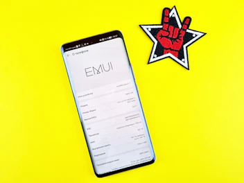 A smartphone displaying its system information screen is placed on a bright yellow surface. To the right of the smartphone, there is a patch or sticker featuring a black and white star outline with a red hand making a rock-and-roll sign in the center.