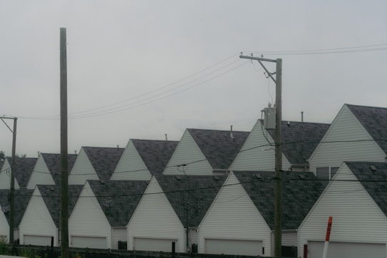 A row of identical houses with triangular roofs, lined up side by side. In the foreground, there are several utility poles with wires stretched across the image, contributing to an urban residential setting. The sky is overcast, casting a muted tone over the scene.