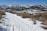 A snowy trail winds through a mountainous landscape, with snow-covered peaks in the distance. The path is bordered by dry grass and low shrubs. The sky is clear and blue.