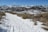 A snowy trail winds through a mountainous landscape, with snow-covered peaks in the distance. The path is bordered by dry grass and low shrubs. The sky is clear and blue.