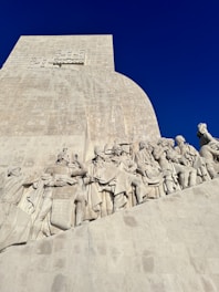 A large stone monument featuring intricate sculptures of historical figures, which appear to be explorers or significant individuals in history. The monument has a smooth, geometric upper section contrasting with the detailed carvings below. It is set against a clear, deep blue sky.