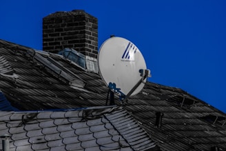 A rooftop with dark shingles and a chimney, featuring a satellite dish mounted prominently. The sky is clear and blue, creating a contrasting background.
