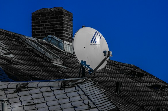 A rooftop with dark shingles and a chimney, featuring a satellite dish mounted prominently. The sky is clear and blue, creating a contrasting background.