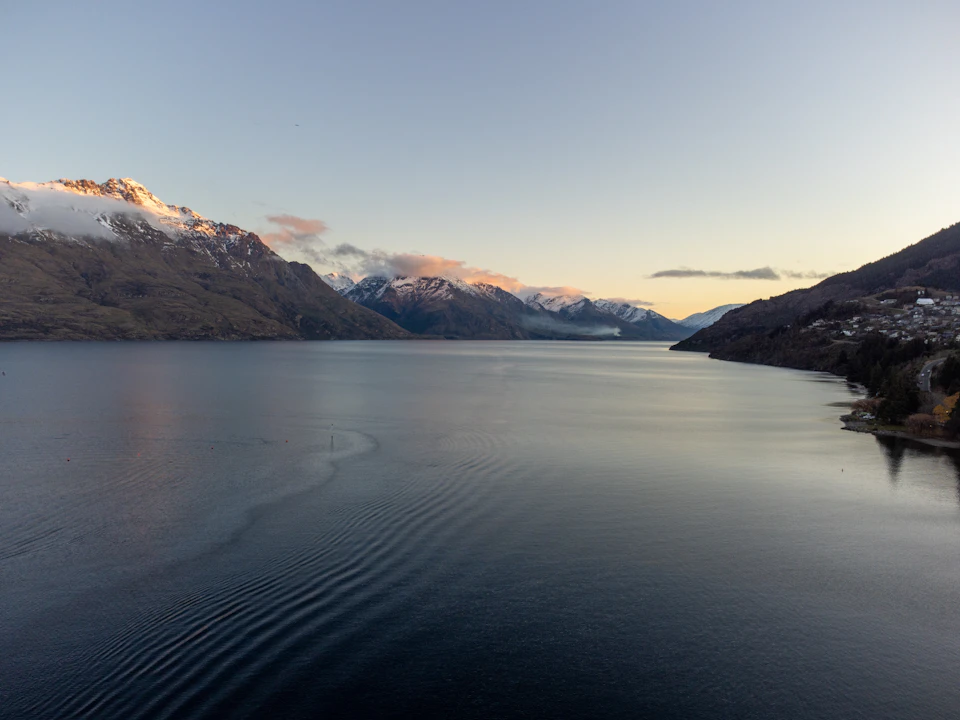 The image depicts a serene mountain lake at sunset, with a light blue sky and snow-capped mountains in the background. The calm water of the lake reflects the warm hues of the setting sun, creating a beautiful contrast between the natural elements. The perspective is from above, providing an aerial view of the landscape below.
