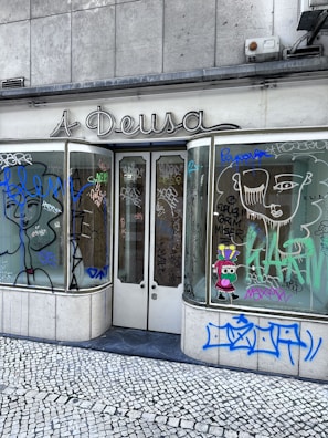 A storefront with graffiti-covered windows and doors, featuring various abstract designs and doodles in multiple colors. The sign above the store reads 'A Deusa'. The ground is covered in cobblestone pavement.