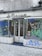 A storefront with graffiti-covered windows and doors, featuring various abstract designs and doodles in multiple colors. The sign above the store reads 'A Deusa'. The ground is covered in cobblestone pavement.