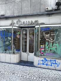 A storefront with graffiti-covered windows and doors, featuring various abstract designs and doodles in multiple colors. The sign above the store reads 'A Deusa'. The ground is covered in cobblestone pavement.