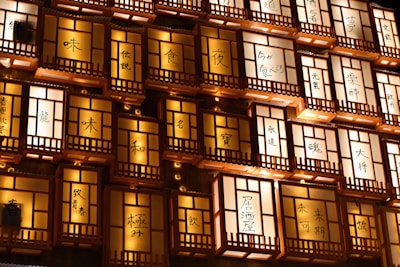Illuminated signs featuring Japanese characters are arranged in a grid-like pattern. The glowing panels are framed with wooden latticework, creating a warm, inviting ambiance. Each panel is uniformly lit, with the characters standing out sharply against the light.